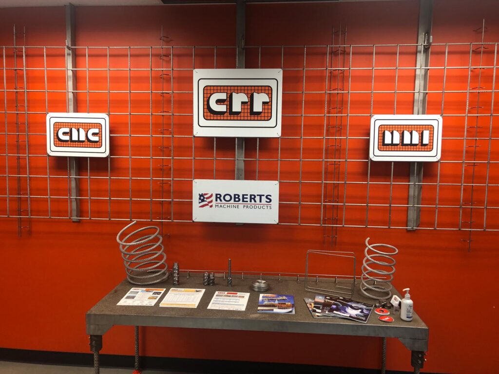 Signs and brochures for CMC, MMI, Roberts Machine Products, and CRR, which are businesses that manufacture the Joint Jockey. The company names are on signs hanging on a wall, while brochures and example products, including two rebar springs, sit on a table below.