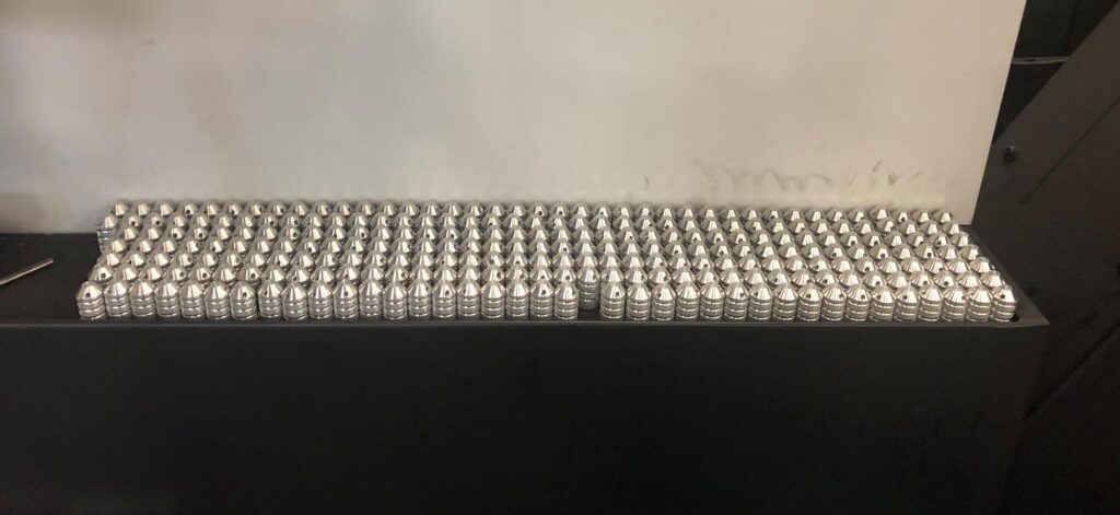 Lots of top caps for the Joint Jockey which have been freshly machined by a skilled CNC machine operator. In this picture dozens of cap sit in a wide rectangle formation, and all the caps are still shiny metallic since the colored-coatings have not yet been added.